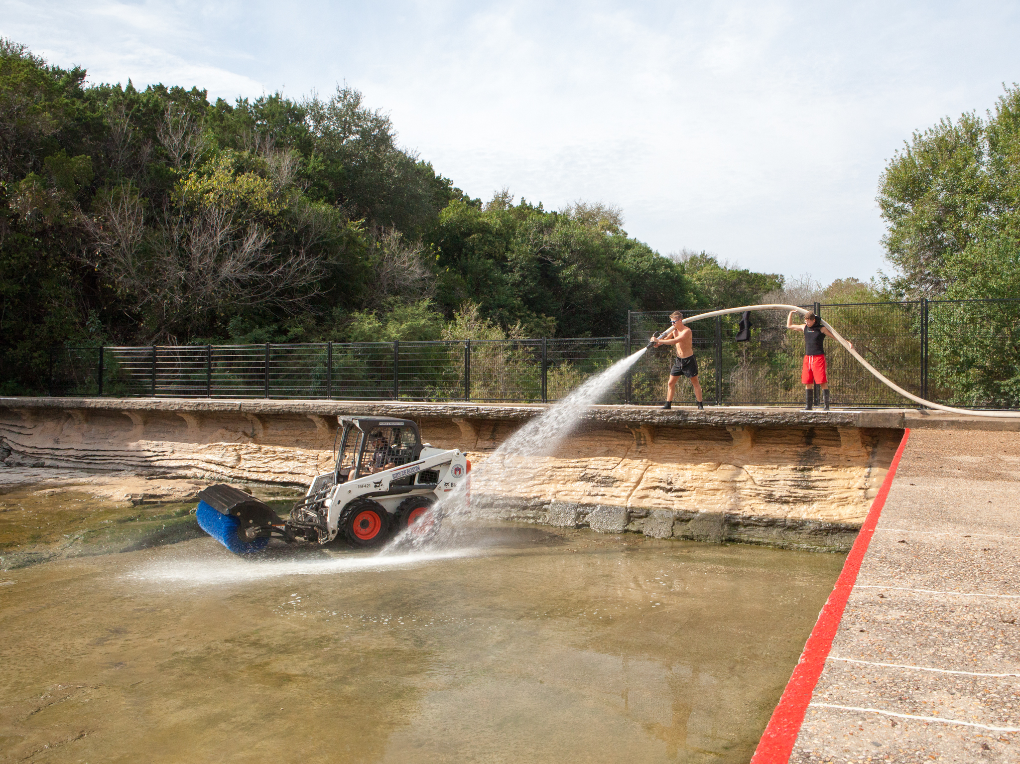 At the shallow end of the pool, where the salamanders don’t live, lifeguards use a firehose and a skidsteer loader equipped with a giant brush to powerwash the pool bottom. The wall they’re standing on blocks the flow of Barton Creek, which would once have flowed through the area where the pool is now. On this day, it’s diverted around the pool into a tunnel beneath the concrete walkway on the right.